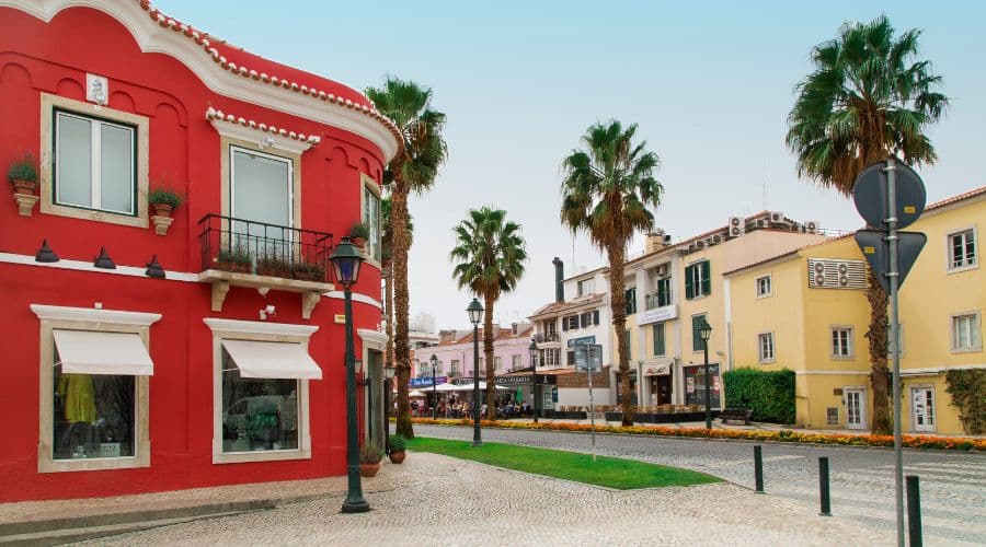 Alameda Combatentes Grande Guerra Cascais. Rua com asfalto vermelho, palmeiras em sua extensão e flores em sua calçada. Casas com cores fortes, amarela e vermelha.