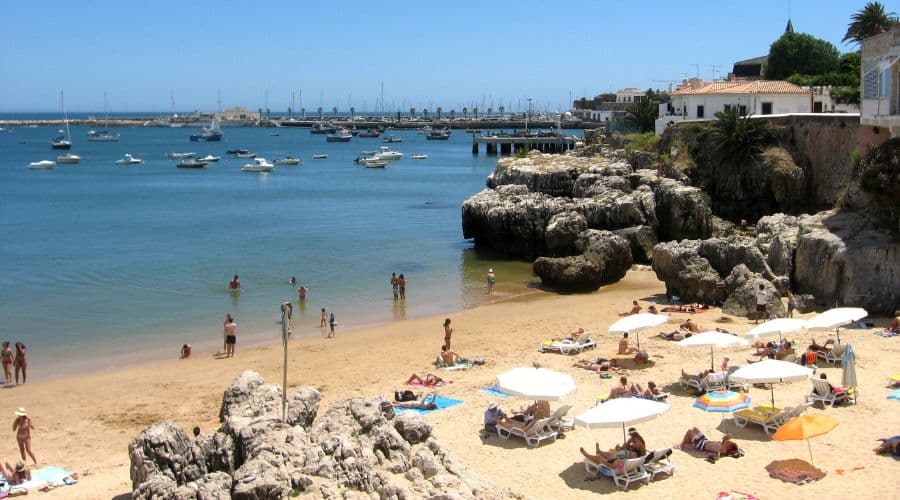 Praia de Cascais. Pessoas tomando bar no mar e guarda sol com algumas pessoas sentadas e deitada na areia