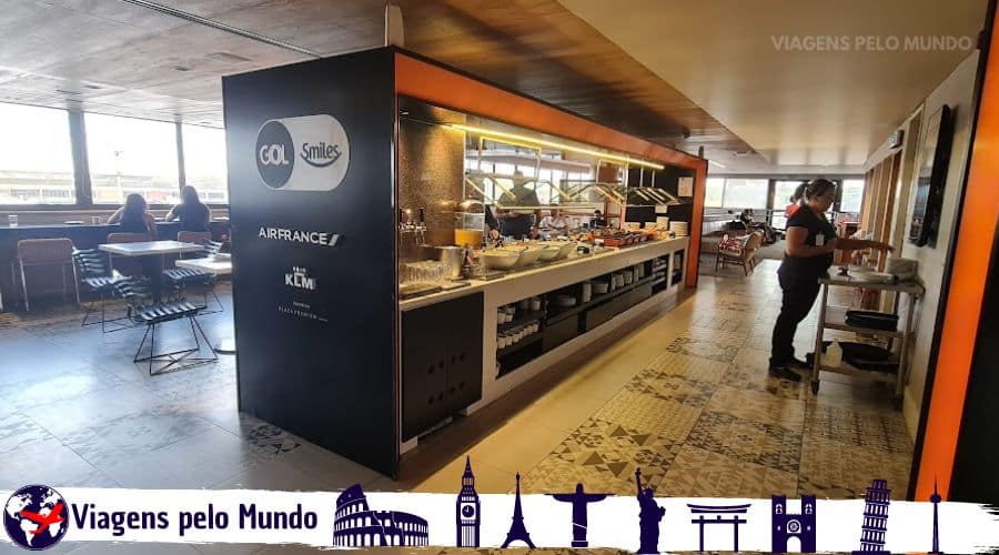 Gol Premium Nacional. Balcão com o buffet com comidas variadas e logo da Gol na parede com pessoas se servindo.