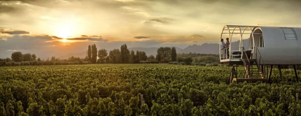 Hotéis vinícola em Mendoza: pôr do sol deslumbrante sobre um vasto vinhedo em Mendoza com há uma estrutura cilíndrica moderna sobre elevação, com uma varanda onde duas pessoas observam o cenário