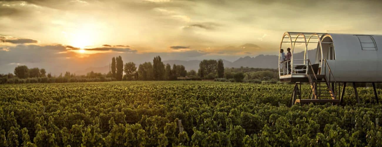 Hotéis vinícola em Mendoza: pôr do sol deslumbrante sobre um vasto vinhedo em Mendoza com há uma estrutura cilíndrica moderna sobre elevação, com uma varanda onde duas pessoas observam o cenário