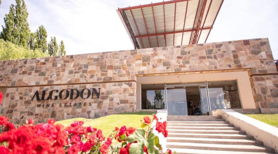 Hotel vinícola em Mendoza: Algodon Wine Estates: entrada destacada por uma imponente parede de pedra com o nome gravado, cercada por flores vermelhas vibrantes e uma estrutura moderna ao fundo. A arquitetura combina elementos rústicos com design contemporâneo, criando um convite visual ao luxo.