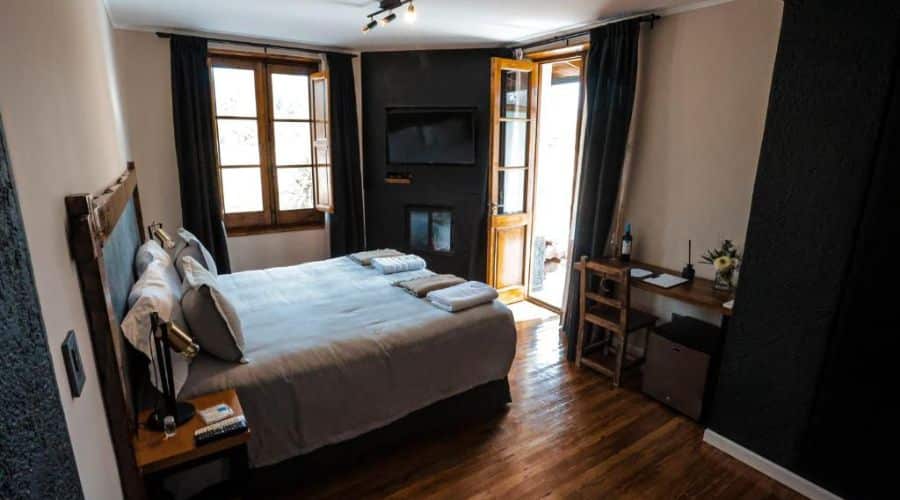 Hotel Algodon Wines Estates: quarto acolhedor dentro da propriedade, com piso de madeira e paredes em tons escuros que contrastam com a cama de cor clara. O ambiente é intimista, com uma lareira que adiciona um toque de conforto e sofisticação. A janela permite a entrada de luz natural, complementando o espaço.