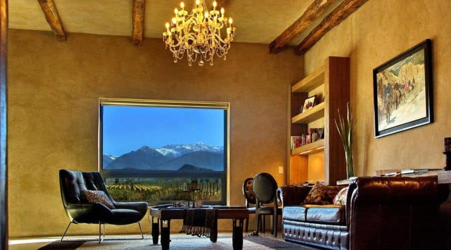 Alpasión Lodge: Sala de estar luxuosa com teto de vigas de madeira, candelabro pendente e uma grande janela com vista para as montanhas. A sala é decorada com sofás de couro, poltronas confortáveis e estantes embutidas, criando um ambiente elegante e aconchegante.