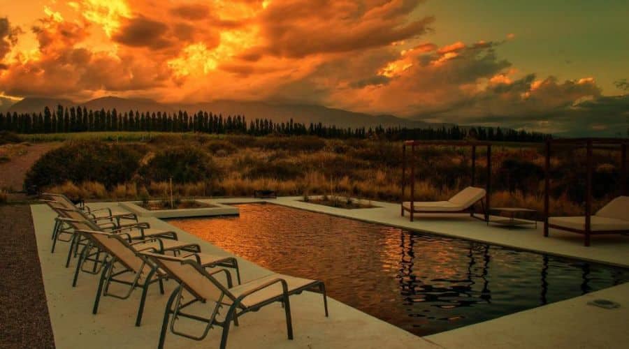 Piscina ao ar livre ao pôr do sol, com espreguiçadeiras dispostas ao redor. O céu está iluminado com tons laranja e dourado, refletindo na água da piscina. A paisagem ao redor é composta por campos e árvores, criando um cenário de pôr do sol.