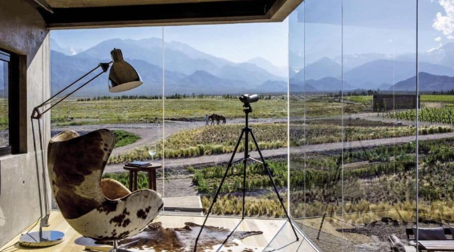 Vale de Uco Resort: Quarto com Vista Ampliada. Destaca uma cadeira moderna ao lado de uma janela de vidro do chão ao teto, oferecendo uma vista espetacular da paisagem natural externa, ideal para contemplação ou leitura