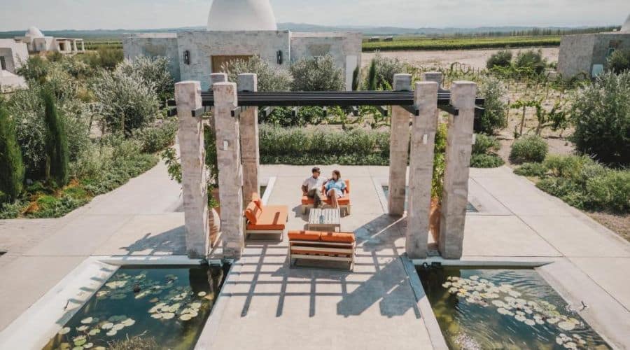 Hotéis vinícolas em Mendoza, Chozos: Área externa do hotel vinícola com mobiliário moderno, incluindo sofás laranja e uma mesa central. O espaço é cercado por colunas de pedra e espelhos d'água com plantas aquáticas. Ao fundo, o edifício principal do hotel e os vinhedos completam o cenário.