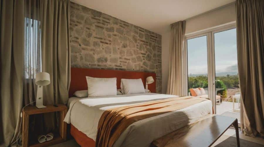 Quarto acolhedor com cama de casal, cabeceira em tecido laranja e parede de pedra. O quarto tem uma porta de vidro que leva a uma varanda, oferecendo vista para a paisagem ao ar livre. A decoração é simples e elegante, com tons neutros e detalhes em madeira.