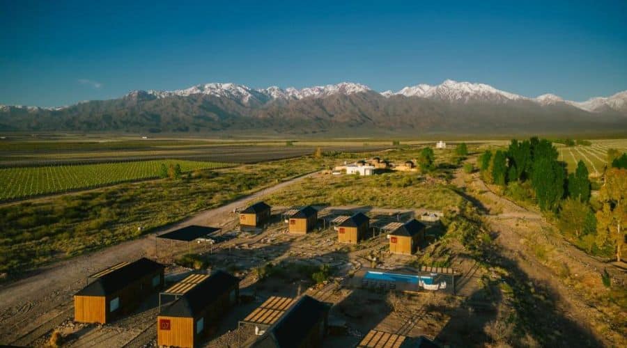 Hotéis vinícolas em Mendoza, La Morada Lodge: Vista aérea de um conjunto de chalés modernos em meio a um vasto vinhedo, com montanhas cobertas de neve ao fundo. O céu está claro, realçando a beleza da paisagem e a disposição harmoniosa dos chalés.