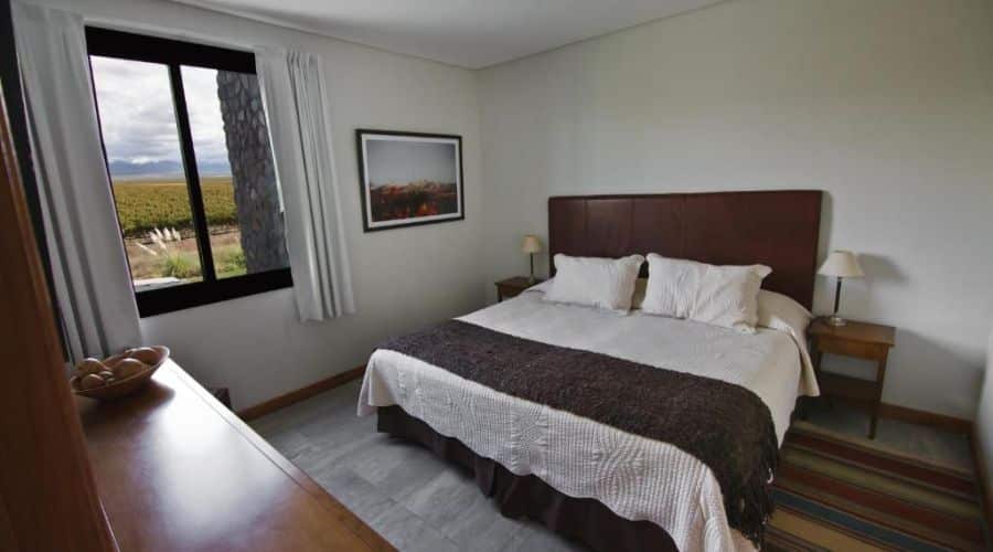 Quarto aconchegante do La Morada Lodge com uma grande cama de casal, decorado em tons neutros. A janela oferece uma vista para os vinhedos, trazendo a natureza para dentro do ambiente. A decoração é simples e elegante, com móveis de madeira e um quadro na parede.