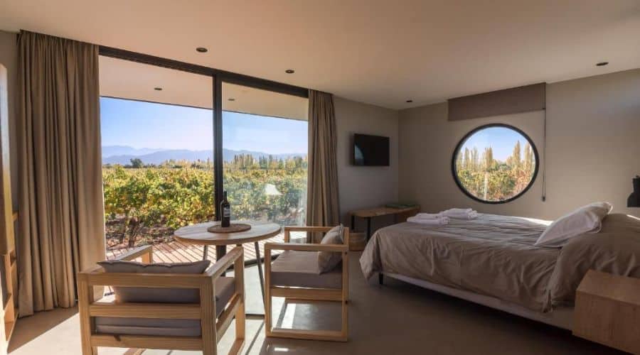 Posada Mawida: Interior de um quarto moderno e aconchegante, com uma grande janela que oferece uma vista panorâmica dos vinhedos e das montanhas ao fundo. O quarto possui uma cama grande, uma mesa redonda com cadeiras e decoração minimalista, proporcionando um ambiente relaxante e sofisticado.
