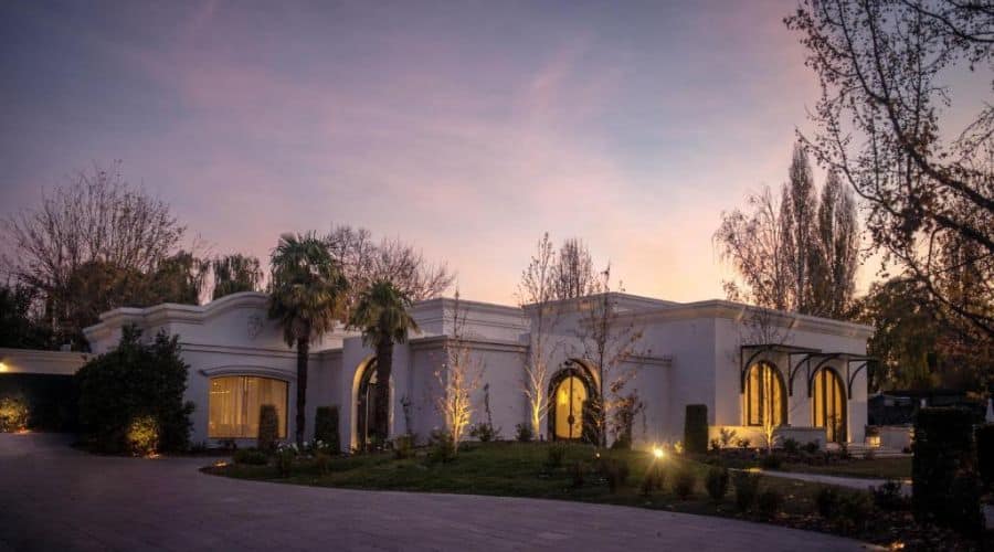 Hospedagem Suzana Balbo: Fachada de uma elegante vinícola ao entardecer, com iluminação suave destacando a arquitetura refinada. O céu ao fundo exibe um tom rosado, adicionando um toque de tranquilidade ao cenário. Árvores e plantas ao redor completam o ambiente sereno.