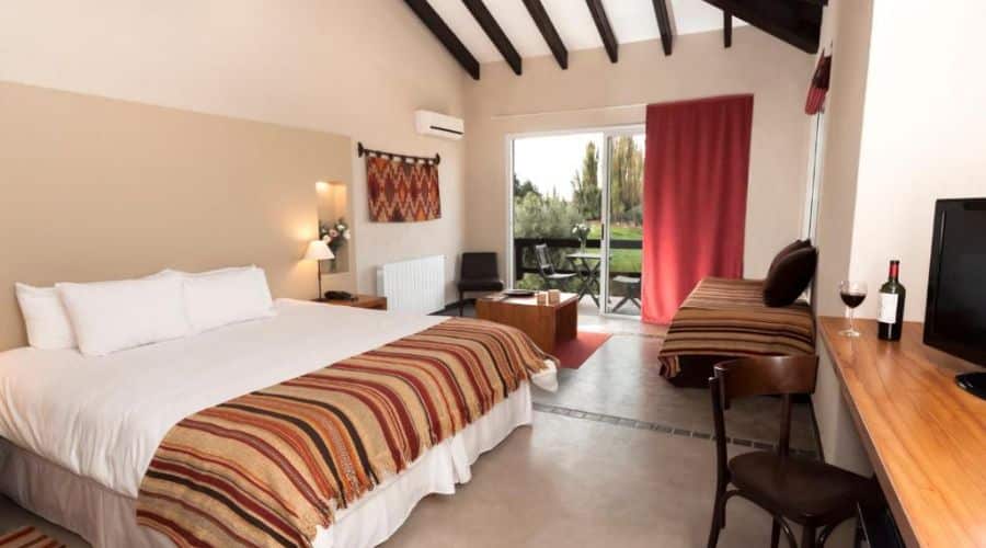 Hotel Villa Mansa Mendoza: quarto espaçoso e bem iluminado com uma grande janela de vidro que se abre para uma varanda com vista para um ambiente com muito verde. Há também uma área de trabalho com uma mesa e cadeira, uma televisão de tela plana, e uma garrafa de vinho com uma taça
