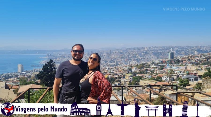 Conhecendo Valparaiso no Chile. Casal (a gente) posando para uma foto em um mirante com vista panorâmica de Valparaíso ao fundo.