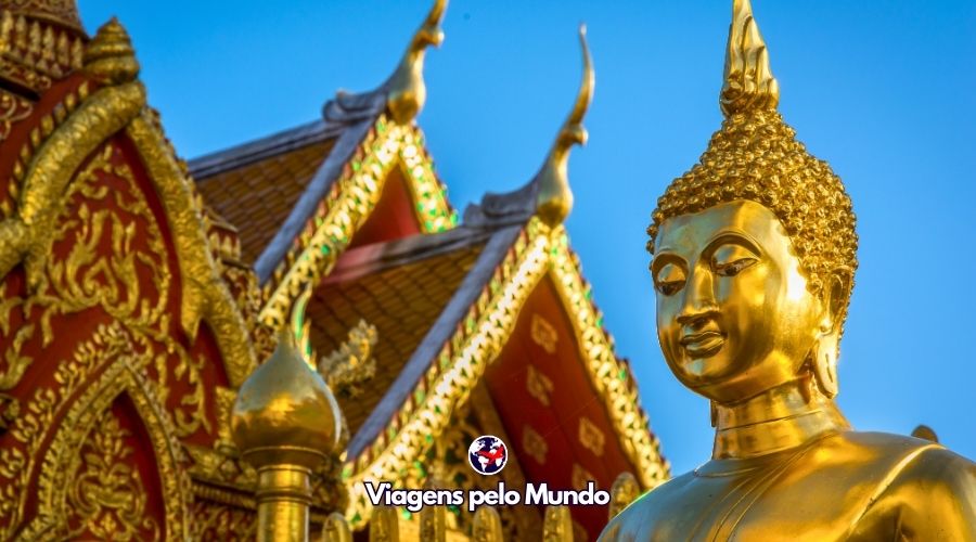 Os templos da Tailândia são uma imersão cultural no país. Imagem do Buda com um templo no fundo.